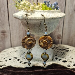May include: A pair of handcrafted earrings featuring brown, fossil-like round beads. The earrings have a bronze-colored hook and are accented with small blue beads and a larger, oval-shaped bead with blue and yellow hues. The earrings hang in front of a white decorative vase.