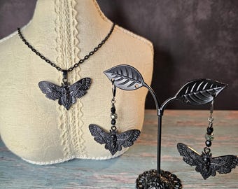 Puede incluir: Conjunto de collar y pendientes de polilla negra. El collar presenta un gran colgante de polilla detallado con cuentas negras. Los pendientes son pendientes colgantes con un pequeño amuleto de polilla y cuentas negras.