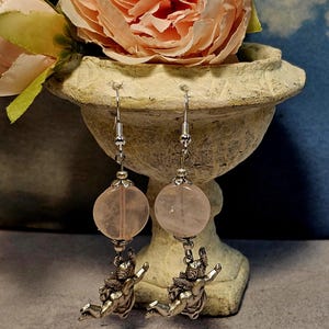 May include: A pair of dangle earrings featuring large, round, pale pink rose quartz beads. Below each bead hangs a silver-toned cherub charm. The earrings are suspended from silver-colored fish hook ear wires. The earrings are displayed in front of a decorative vase.