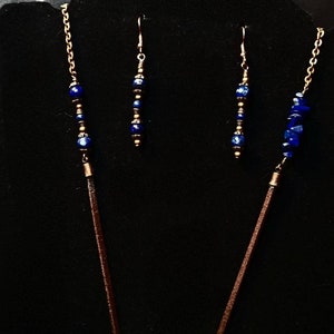 Op de afbeelding: Een set sieraden met een lange ketting met een bruin leren koord en een ketting met blauwe lapis lazuli kralen, en bijpassende oorbellen met blauwe lapis lazuli kralen en gouden accenten.