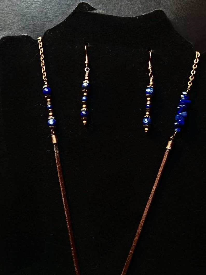 Op de afbeelding: Een set sieraden met een lange ketting met een bruin leren koord en een ketting met blauwe lapis lazuli kralen, en bijpassende oorbellen met blauwe lapis lazuli kralen en gouden accenten.