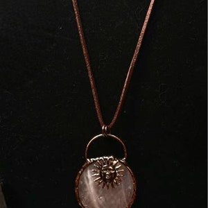 Op de afbeelding: Een ketting met een hanger van rozenkwarts met een zonnengezichtontwerp en een bedel van helder kwartskristal. De ketting is gemaakt van bruin lederkoord en goudkleurige metalen accenten. De oorbellen zijn gemaakt van blauwe lapis lazuli kralen en goudkleurig metaal.