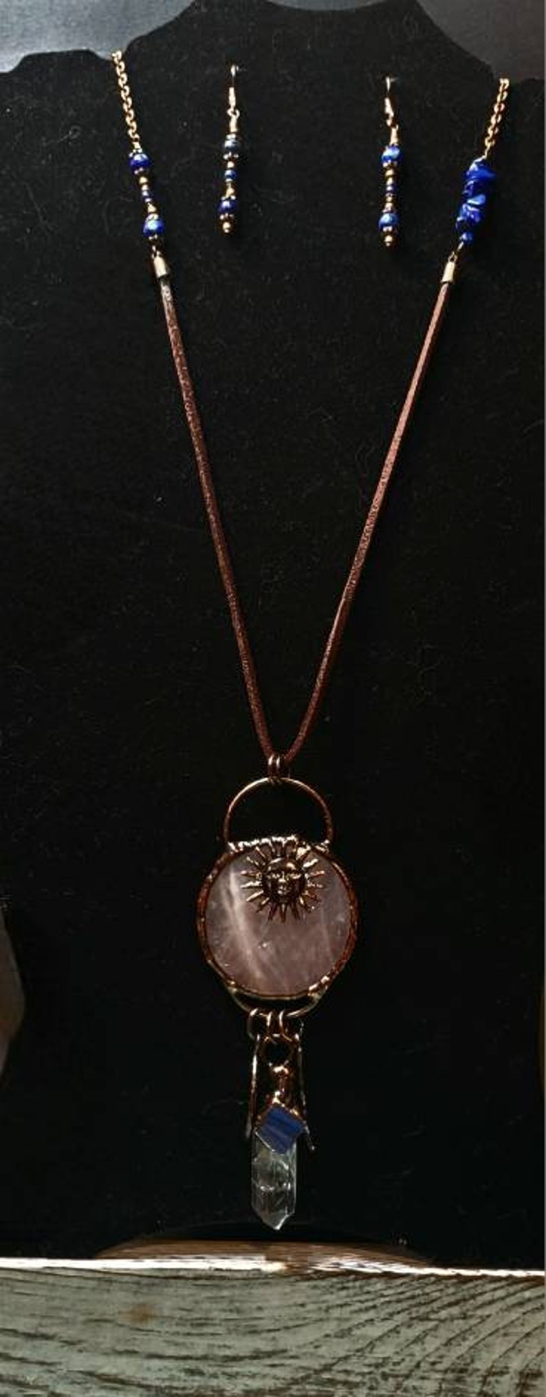 Op de afbeelding: Een ketting met een hanger van rozenkwarts met een zonnengezichtontwerp en een bedel van helder kwartskristal. De ketting is gemaakt van bruin lederkoord en goudkleurige metalen accenten. De oorbellen zijn gemaakt van blauwe lapis lazuli kralen en goudkleurig metaal.