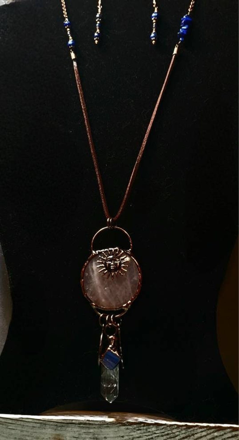 Op de afbeelding: Een koperen ketting met een hanger van rozenkwarts met een zonontwerp. De hanger is bevestigd aan een lang bruin leren koord met blauwe lapis lazuli kralen. De ketting bevat ook bijpassende oorbellen.
