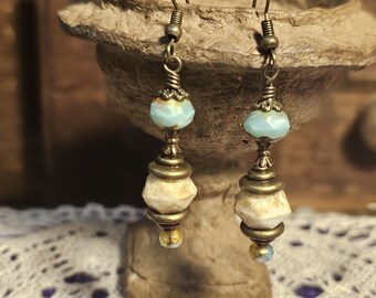 May include: A pair of handcrafted earrings with a vintage aesthetic. Each earring features a brass-colored hook, a light blue bead, and a stacked design of cream-colored and brass-toned beads. The earrings are displayed against a textured background.