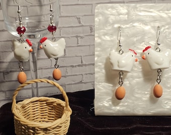 Pode incluir: Brincos com tema de galinha com figuras de galinha brancas, detalhes de crista vermelha e contas em forma de ovo. Os brincos são exibidos com uma pequena cesta de vime e um copo.