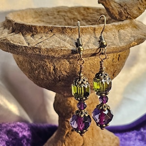 May include: A pair of dangle earrings with green and purple glass beads. The earrings have a bronze finish and are hanging from a bird bath.