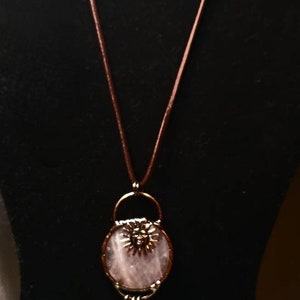 Op de afbeelding: Een ketting met een hanger van roze rozenkwarts met een koperen zonontwerp en een hanger van helder kwartskristal. De ketting is aan een bruin leren koord met blauwe glazen kralen bevestigd.