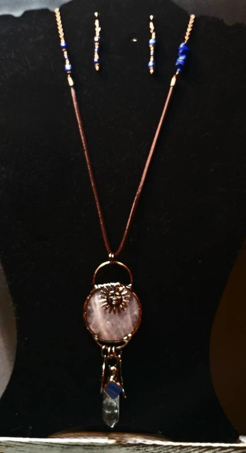 Op de afbeelding: Een ketting met een hanger van roze rozenkwarts met een koperen zonontwerp en een hanger van helder kwartskristal. De ketting is aan een bruin leren koord met blauwe glazen kralen bevestigd.