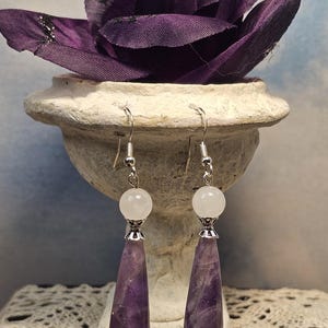 May include: A pair of dangle earrings featuring a white, round bead at the top, a silver-colored metal accent, and a teardrop-shaped, purple amethyst stone. The earrings are displayed in front of a purple flower and a decorative vase.