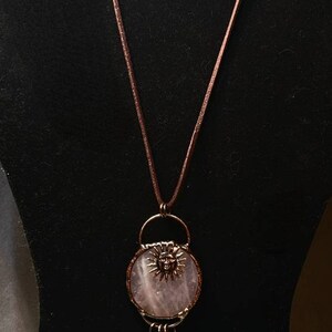 Op de afbeelding: Een ketting met een hanger van rozenkwarts met een zonnengezicht, een helder kwartskristal en blauwe lapis lazuli accenten. De ketting is aan een bruin leren koord.
