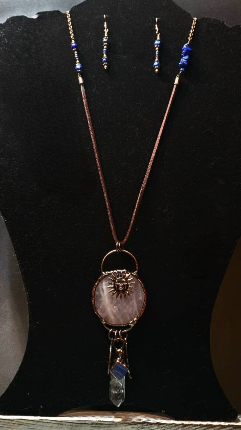 Op de afbeelding: Een ketting met een hanger van rozenkwarts met een zonnengezicht, een helder kwartskristal en blauwe lapis lazuli accenten. De ketting is aan een bruin leren koord.
