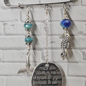 May include: A silver safety pin with dangling charms. Charms include a whale tail, a seahorse, and a circular pendant with the text "may you always have a shell in your pocket & sand between your toes." The charms have blue and silver beads.