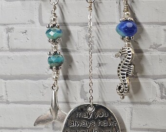 May include: A silver safety pin with dangling charms. Charms include a whale tail, a seahorse, and a circular pendant with the text "may you always have a shell in your pocket & sand between your toes." The charms have blue and silver beads.