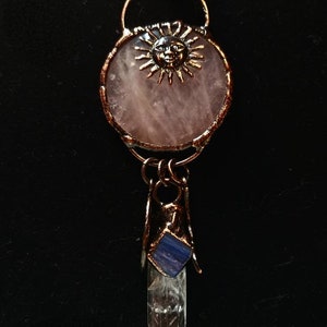 Op de afbeelding: Een ketting met een hanger van koperdraad gewikkeld rond een rozenkwarts steen en een zon gezicht charme. Een helder kwarts kristal met een blauwe steen is bevestigd aan de onderkant van de hanger.