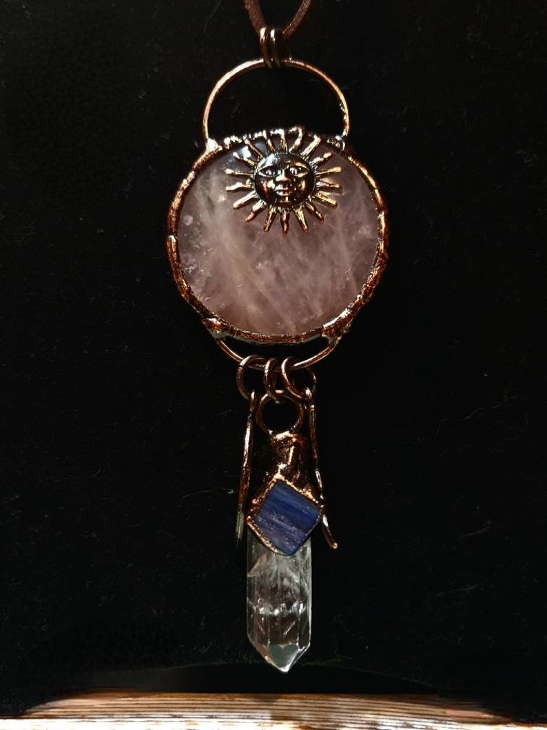Op de afbeelding: Een ketting met een hanger van koperdraad gewikkeld rond een rozenkwarts steen en een zon gezicht charme. Een helder kwarts kristal met een blauwe steen is bevestigd aan de onderkant van de hanger.