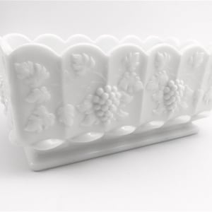 May include: A white rectangular planter with a scalloped rim and a raised base. The planter features embossed grape and leaf designs on the sides. The overall design is a classic, vintage style.
