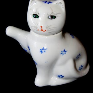 May include: A white ceramic cat figurine with a raised paw, featuring small blue floral patterns across its body. The cat has green eyes, a small orange nose, and delicate grey whiskers painted on its face, with a blue flower on its forehead.
