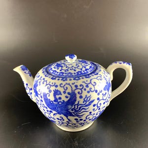 May include: A white ceramic teapot with a blue phoenix design and matching lid. The teapot has a curved spout and handle, and the design includes floral and swirl patterns. The teapot is a traditional style.