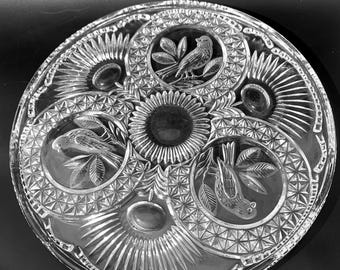 May include: A clear glass cake plate with a decorative design of birds and leaves. The plate has a scalloped edge and a raised center.