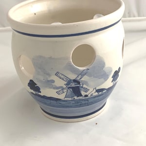 May include: A white ceramic garlic keeper with a blue windmill and landscape design. The keeper has three round holes and a blue rim. The design is reminiscent of Delftware pottery.
