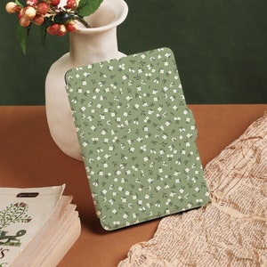 Könnte beinhalten: Eine E-Reader-Hülle mit einem grün-weißen Blumenmuster. Die Hülle liegt auf einer braunen Oberfläche mit einem zerknitterten Stück Papier und einer weißen Vase mit roten Beeren.