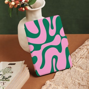 Könnte beinhalten: Eine E-Reader-Hülle mit einem abstrakten Muster in Pink und Grün. Die Hülle liegt auf einer braunen Oberfläche mit einem Stapel Bücher und einer weißen Vase mit roten Beeren.