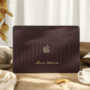 May include: A dark brown laptop case with a textured, crocodile-like pattern. The Apple logo is visible, along with the name "Mark Aldrich" in gold lettering. The case is on a burlap surface, with a cup of coffee and a book in the background.