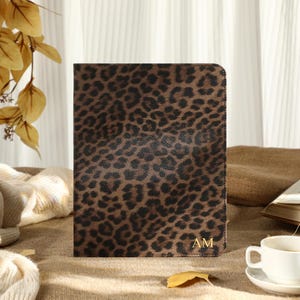 Può includere: Un diario rettangolare con stampa leopardata marrone e nera con le iniziali dorate "AM". Il diario è esposto in posizione verticale su una superficie neutra, con una tazza e un piattino bianchi e un libro aperto. Foglie decorative sullo sfondo.