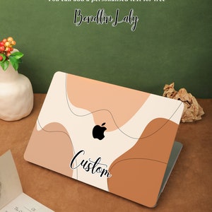 May include: A laptop with a custom design featuring abstract shapes in shades of brown, beige, and white. The design includes the word "Custom" in black cursive lettering. The laptop is on a brown surface with a book and a vase of red berries.