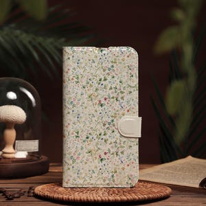 Könnte beinhalten: Eine Handyhülle mit floralem Muster, cremefarbenem Hintergrund und weißem Verschluss. Die Hülle zeigt ein zartes Design aus kleinen Blumen und Blättern in verschiedenen Farben. Die Hülle liegt auf einem gewebten Untersetzer.