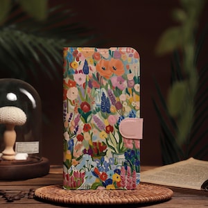 Könnte beinhalten: Eine Handyhülle mit floralem Muster und einem rosafarbenen Verschluss. Die Hülle zeigt ein farbenfrohes Design verschiedener Blumen in Rot-, Orange-, Rosa-, Blau- und Lilatönen. Sie liegt auf einem gewebten Untersetzer auf einer Holzoberfläche.
