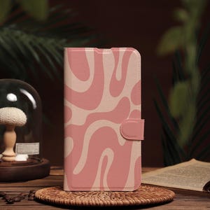 Puede incluir: Una funda de teléfono rosa con un patrón abstracto de espiral blanco. La funda tiene una solapa que se cierra con un botón.