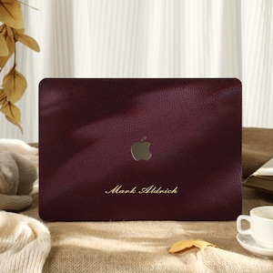 Op de afbeelding: Een bordeauxrode laptophoes met een textuur die op leer lijkt. De hoes heeft een goudkleurig Apple-logo en de naam "Mark Aldrich" in gouden letters. Een witte theekop en schotel staan in de buurt.