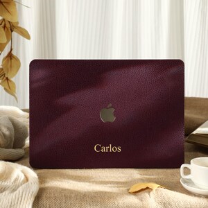 Könnte beinhalten: Eine weinrote Laptop-Hülle mit strukturierter Oberfläche und goldenem Apple-Logo. Der Name "Carlos" ist in goldener Schrift unter dem Logo aufgedruckt. Rechts befinden sich eine weiße Tasse und Untertasse, im Hintergrund ein offenes Buch.