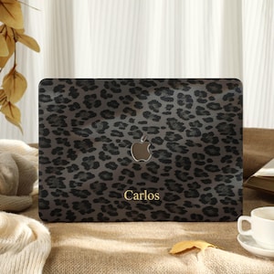 Puede incluir: Una funda para portátil con un diseño de estampado de leopardo en tonos grises y negros. La funda presenta el logotipo de Apple y el nombre "Carlos" en letras doradas. La funda está sobre una superficie marrón.
