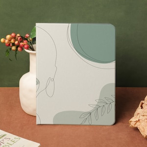 Könnte beinhalten: Ein weißes Notizbuch mit minimalistischem Design, das eine Linienzeichnung eines Gesichts, einen grünen Kreis und einen grünen Zweig mit Blättern zeigt. Das Notizbuch hat eine strukturierte Oberfläche.