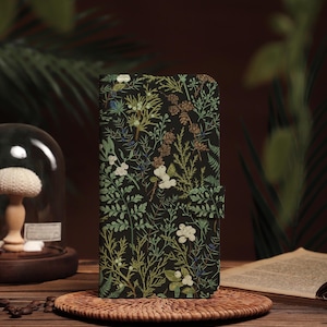 Könnte beinhalten: Eine schwarze Handyhülle mit einem floralen Muster aus weißen Blüten, grünen Blättern und braunen Akzenten. Die Hülle mit Magnetverschluss wird auf einem gewebten Untersetzer präsentiert, was ein naturinspiriertes Thema suggeriert.