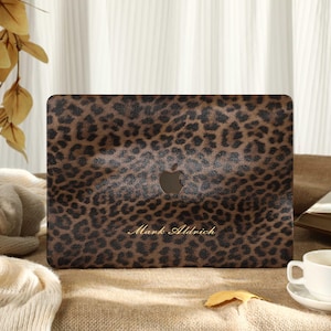 May include: A laptop case with a brown and black leopard print design. The case has the Apple logo and the name "Mark Aldrich" in gold lettering. The case is on a brown surface with a white cup and saucer.