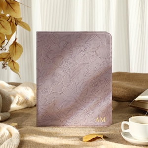 Può includere: Una custodia per tablet lilla con un motivo floreale in rilievo. Le iniziali "AM" sono dorate in basso a destra. Una tazza e un piattino bianchi sono davanti. Lo sfondo include un libro, una coperta beige e foglie decorative.