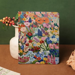 May include: A notebook cover with a bright floral design, showcasing a variety of flowers in orange, pink, blue, and purple hues. The rectangular cover is likely made of a smooth material. A white vase with berries is to the left.