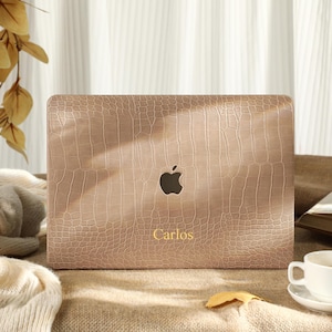 Può includere: Una custodia per laptop beige con una texture a pelle di coccodrillo. Il logo Apple è visibile e il nome "Carlos" è stampato in oro. Una tazza e un piattino bianchi sono su un tavolo con un libro e foglie autunnali.