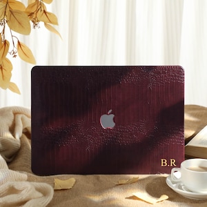 May include: A burgundy laptop case with a textured, embossed pattern. The case features a silver apple logo and gold initials "B.R." A white teacup and saucer sit nearby. The background includes a book and decorative leaves.