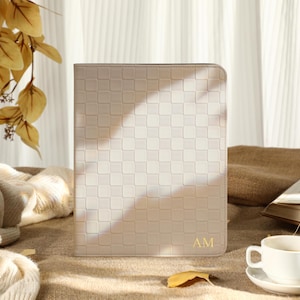 Può includere: Una custodia beige per tablet con un motivo a quadri e le iniziali dorate "AM". La custodia è su un tavolo con una tazza di caffè, un libro e un maglione color crema.