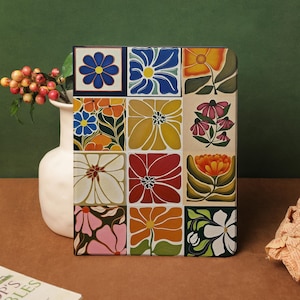 Puede incluir: Un cuaderno cuadrado con un estampado floral que presenta nueve diseños de flores coloridas. Las flores son en tonos de azul, naranja, amarillo, rojo y rosa. Un jarrón blanco con bayas y un libro están en el fondo.