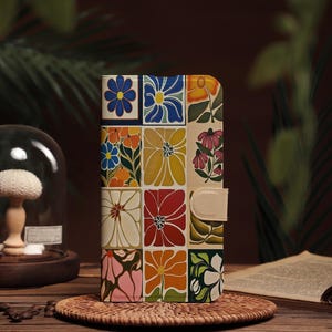 Könnte beinhalten: Eine Handyhülle mit floralem Muster, die ein Raster aus bunten Blumen in Blau, Orange, Gelb, Rot und Rosa zeigt. Sie hat einen Verschluss mit beigem Lederriemen. Die Hülle wird auf einem gewebten Untersetzer präsentiert.