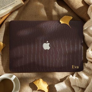 Può includere: Una custodia per laptop in pelle marrone scuro con motivo strutturato, con il logo Apple e il nome "Eva" in lettere dorate. L'immagine include una tazza di caffè, un libro aperto e foglie autunnali.
