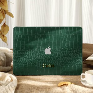 Könnte beinhalten: Eine dunkelgrüne Laptop-Hülle mit Krokodilhaut-Textur. Das Apple-Logo ist sichtbar, und der Name "Carlos" ist in Gold gedruckt. Die Hülle befindet sich auf einer braunen Oberfläche, mit einer weißen Tasse und Untertasse im Vordergrund.