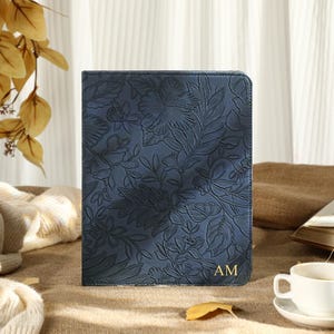 May include: A navy blue journal with a floral embossed design and gold initials. The journal is standing upright on a neutral-colored surface. A white teacup and saucer are in the foreground, with a book and decorative leaves in the background.