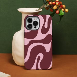 Peut inclure: Une coque de smartphone avec un motif tourbillonnant dans des tons de rose et de bordeaux. L'étui est présenté à côté d'un vase blanc avec des baies et des feuilles. La coque du téléphone possède un module caméra à trois objectifs.